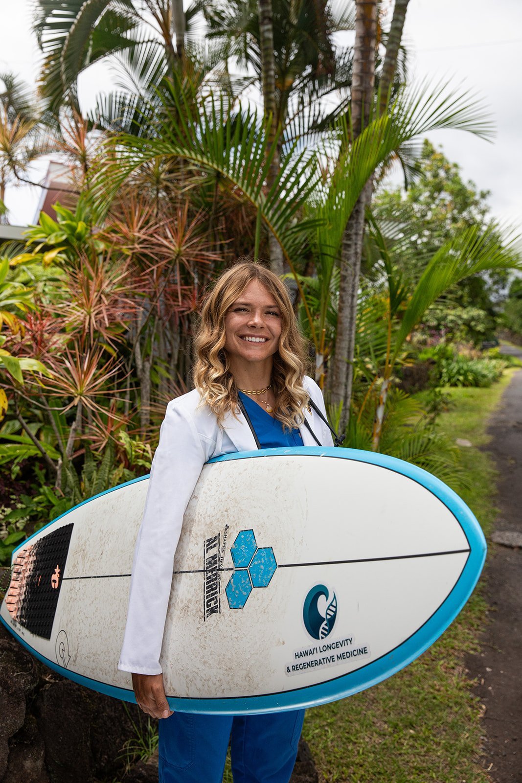 Woman in white blazer holding surfboard with blue trim stands smiling in tropical garden setting with palm trees